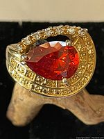 Front view of pear-shaped orange faceted stone ring with clear accent stones and gold-tone ornate band, showing gemstone brilliance and decorative detailing.