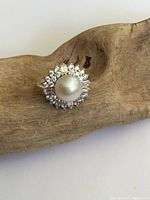 Close-up photo of the ring showing the central round pearl and surrounding clear crystals on a wooden background.