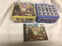 Photo of two metal tins with Beatles album artwork and a sealed CD of 'Sgt. Pepper's Lonely Hearts Club Band' in front.