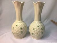 Two cream-colored Belleek bud vases with raised woven basket texture and green shamrock decorations, standing side by side against a white cloth background