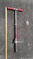Red pogo stick with red foam handles, metal tube body, and rusted spring base placed on asphalt surface alongside a ruler.