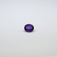 Clear image of the faceted oval cut amethyst gemstone lying on a white surface showing its purple color and size.