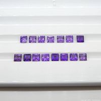 Fifteen square-cut amethyst gemstones displayed in two rows on a white grooved surface, showing their size, shape, and color.