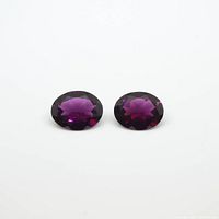 Pair of oval cut garnet gemstones placed side by side on a white background, clearly showing the deep purple color and faceting.