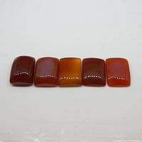 Top-down view of five rectangular cabochon carnelian gemstones laid in a line on a white background, showing red to orange coloration and glossy finish.