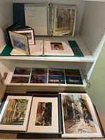 Photo of photo albums on shelves with some prints and photographs visible in plastic sleeves and photo mounts showing urban and landscape scenes.