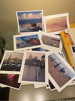 Assorted landscape and people photographic prints laid out on a surface, showing various scenes including clouds reflected on water, sunset, buildings, and flowers.