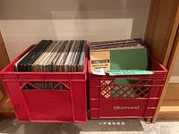 Two red crates filled with assorted vinyl records, viewed from the front showing various album spines and covers