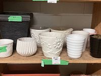 Shelf view showing multiple pots including large square black plastic pot, white ceramic pots with floral embossing and ribbed texture