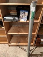 Wide view of wooden shelf holding Epson Powerlite 76c projector, quick setup manual, and nearby tripod projection screen in metallic tube.
