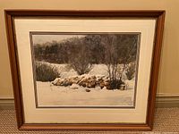 Full view of framed watercolor painting showing winter landscape with snow, trees, and woodpile.