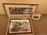 Three framed artworks displayed against a wall on carpet: a large batik market scene, a medium landscape painting, and a small landscape painting.