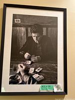 Framed black and white photograph showing a male figure playing cards, with cards spread on a rough wooden table. The man is dressed in a dark jacket and smoking a cigarette. The frame is black with a white matte, measuring 18.5 by 25 inches.