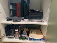 Shelf with assorted office supplies including multiple binders, CD cases, three-hole punch, Dust Off cans, and file folder labels.