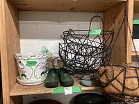 Photo of wooden shelf with large black wire hanging planter, white ceramic planter with floral/bird design, green garden clogs, small stone bird figurine, and butterfly garden stake.