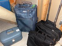 Roots navy blue roller suitcase, two black computer bags, and vintage blue vanity case shown grouped on floor.