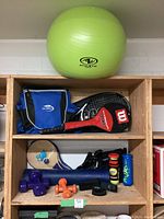 Green Athletic brand exercise ball, US Open backpack, Wilson Titanium badminton racket and cover, Prince tennis racket and cover, ping pong paddles and ball, tennis balls, blue yoga mat, and various dumbbells on a wooden shelf
