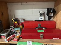Two red metal Mastercraft toolboxes, one large and one small, with various tubes and hardware items arranged behind and around them on a shelf. A black plastic funnel is visible behind the toolboxes.