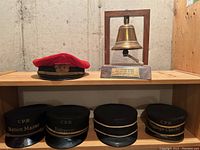 One red CPR conductor-style hat, four black CPR role hats on lower shelf, and brass bell mounted on wooden frame on upper shelf