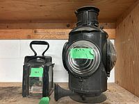 Front and side view of two railroad collectibles on wood shelf: large CNR light with round glass lenses and black metal body, smaller rectangular black metal lantern with glass panes and handle.