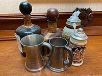 Photo showing two vintage liquor decanters with round stoppers, two pewter tankards in front on wooden surface.