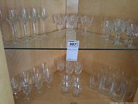 Wide view of upper and lower shelves showing a variety of stem glasses and liquor glasses arranged on two glass shelves against a wooden cabinet interior.