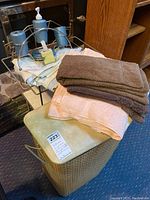 Photo shows folded brown and peach towels stacked on top of a tan woven hamper with a cushioned seat lid; blue dispenser and matching bathroom accessories arranged on a silver metal rack in the background.