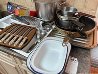 Photo showing large stock pot, frying pans, enamel baking dish, knife holder with knives, measuring cups, grater, and kitchen wrap/foil boxes.