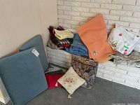 Overall lot showing 6 cushions of different styles and colors, assorted blankets including plaid, orange, and blue blankets, and vinyl tablecloths folded underneath.
