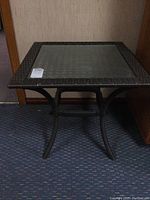 Full view of square patio side table with glass top and dark brown wicker-style frame.