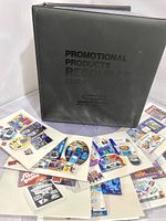 Gray binder labeled 'Promotional Products Resource Guide' shown with multiple promotional photos spread out to show variety.