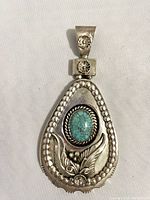 Front close-up of teardrop-shaped sterling silver pendant with turquoise stone and detailed silver work with floral and leaf designs.