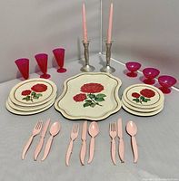 Full display of vintage doll dish set with rose-patterned metal plates, pink plastic silverware, pink plastic glasses and ice cream dishes, metal tray, and metal candlestick holders with pink plastic candles.