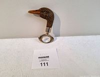 Photo of vintage Comoys of London duck head bottle opener standing on table against wall, showing side view