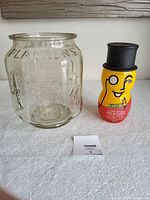 Clear side embossed glass store counter display jar next to Mr. Peanut shaped collectible jar with black top hat lid and yellow inner lid on white surface.