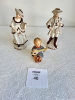 Front view of three figurines: Victorian style man and woman, and seated Hummel child figurine with musical instrument.