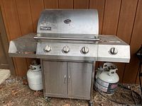 Full front view of large stainless steel Charmglow barbecue grill with three control knobs, storage cabinet below, and two propane tanks, one positioned under the left side shelf, and one beside the unit on right.