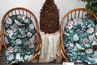 Pair of bamboo swivel chairs with green floral and bird pattern cushions on either side of a small table with a brown artificial tree.