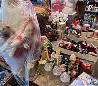 Wide view of Christmas decorations including plush Santa with bears, multiple figurines, glass jars, candle holders, and mugs on a table in a garage setting.