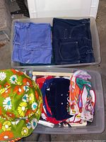 Two plastic bins, one with folded blue denim jeans including Wrangler brand, the other with floral hats and various baby clothes.