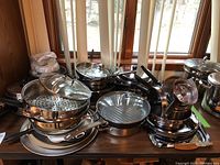 Wide view of various stainless steel pots, pans, lids, steamer, and pizza trays arranged on a table.