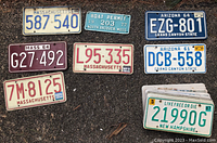 Photo showing a group of license plates spread on pavement including Massachusetts, Arizona, and New Hampshire plates, plus a boat permit plate.