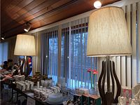 Pair of lamps on display on table among other estate sale items, showing overall appearance and scale next to window blinds.