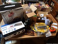 Overview photo of lot items including boxed tools, security devices, binoculars, tape measures, and glasses on a table.