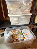 Three stacked plastic storage totes in a garage holding folded vintage sheets with floral patterns and assorted colors.