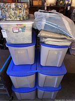 Seven large plastic totes stacked with blue lids, some clear totes with yarn visible inside, and two tote labels visible with yarn color and weight details.