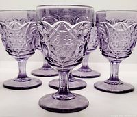 Front view of 6 lavender colored pressed glass goblets arranged closely showing detailed pressed patterns.