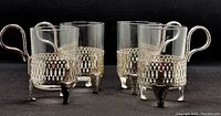 Four clear cylindrical glass bar glasses each set in silver tone metal open lattice style holders with three clawed legs and a handle.