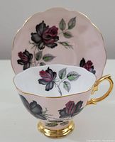 Tea cup and saucer with pastel blush pink background and black and dark pink rose floral design and gold trim on edges and handle.