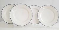Four white ceramic plates with scalloped edge and black stripe, seen front view, standing on clear display stands.